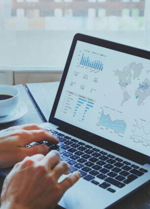 Eine Person hat die Hände auf der Tastatur eines ausgeklappten Laptops, neben dem eine Tasse Kaffee steht. Der Bildschirm zeigt eine Weltkarte und Kursverläufe.