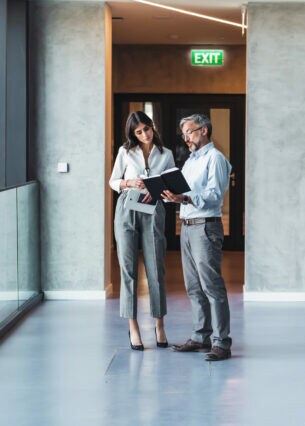 Ein Mann und eine Frau in Business-Kleidung stehen in einem hellen Flur. Er hält ein geöffnetes Buch in einer Hand, in das beide hineinschauen.