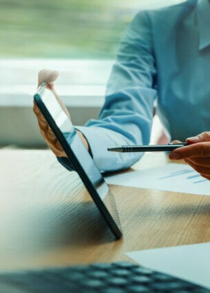 Zwei Personen in Businesskleidung am Schreibtisch, eine Person hält ein Tablet, auf das die andere deutet.