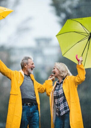 Zwei ältere Personen in gelben Regenmänteln halten im Regen ihre gelben Regenschirme in die Höhe und schauen sich lachend an.