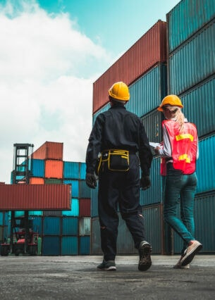 Zwei Personen in Arbeitskleidung und gelben Schutzhelmen gehen neben gestapelten Containern entlang. Im Hintergrund hebt ein Kran einen Container.