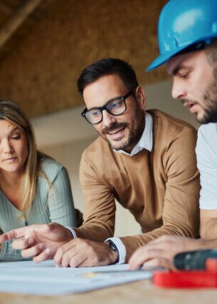 Drei Personen stehen gebeugt über Unterlagen in einem Rohbau.