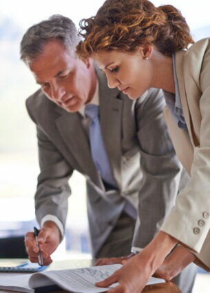 Eine Frau und ein Mann in Businesskleidung stehen über einen Tisch gebeugt, auf dem sich gebundene und aufgeschlagene Papierunterlagen befinden, auf die der Mann mit einem Stift deutet.