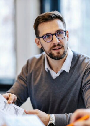 Eine Person hält ein Dokument mit Diagrammen in der Hand und schaut eine Person an, die ihm gegenübersitzt.