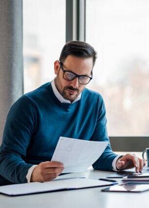 Eine Person sitzt an einem Schreibtisch, schaut in Unterlagen und tippt etwas in einen Laptop.