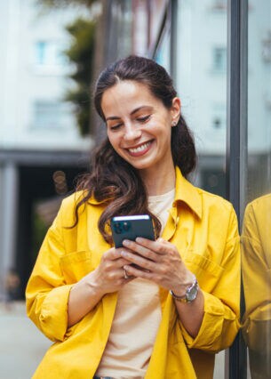 Eine Person lehnt sich mit der linken Schulter an eine Glasfassade und schaut lächelnd auf ihr Smartphone, das sie in den Händen hält.