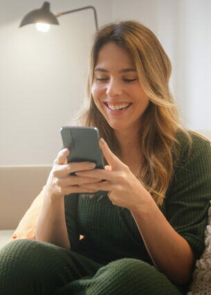 Eine Person sitzt auf einem Sofa und schaut lachend auf das Smartphone, das sie mit beiden Händen festhält.