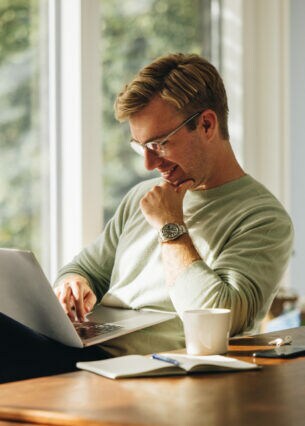 Eine Person sitzt am Esstisch und hat einen geöffneten Laptop auf dem Schoß.