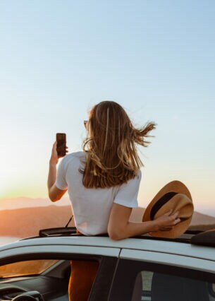 Eine Person in der Rückansicht schaut bei Sonnenuntergang aus dem Dach eines Autos und fotografiert sich dabei.