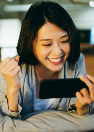 Eine Person liegt auf einem Bett und schaut lächelnd auf das Smartphone, das sie in einer Hand hält.