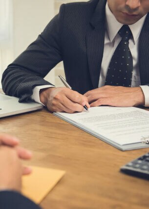 Ein Mann sitzt an einem Tisch vor einem Laptop und unterschreibt ein Dokument. Ihm gegenüber sitzt eine weitere Person.