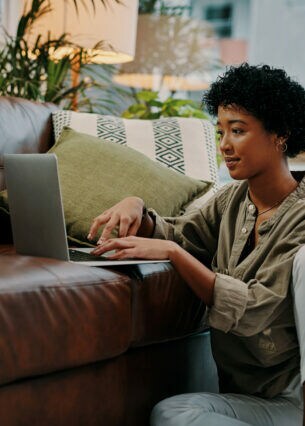 Eine junge Frau sitzt auf dem Boden neben dem Sofa und benutzt ein Notebook.