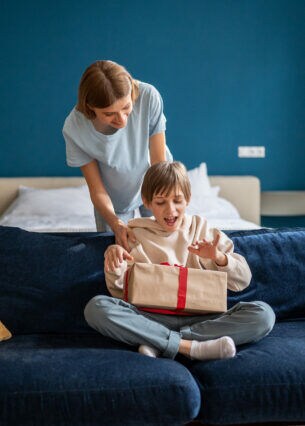 Eine Frau beugt sich über die Rückenlehne eines Sofas, auf der ein Kind sitzt, das ein Paket mit einer roten Schleife auf dem Schoß hat.