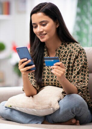 Eine Person sitzt im Schneidersitzt auf einem Sofa und hält sowohl eine Kreditkarte als auch ein Smartphone in den Händen.