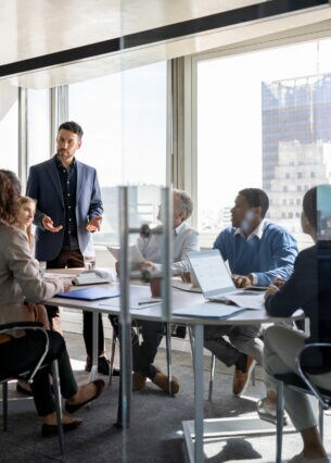 Mehrere Menschen sitzen an einem Tisch in einem Raum mit vielen Fenstern, eine Person steht am Tisch.