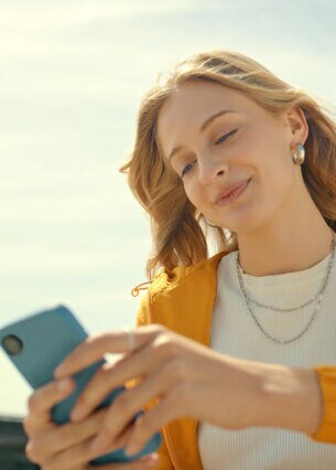 Eine junge Person sitzt im Freien und hält ein Smartphone in ihren Händen.