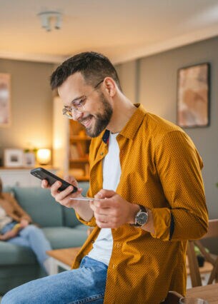 Eine Person sitzt auf einem Tisch und hält eine Kreditkarte sowie ein Smartphone in den Händen. Im Hintergrund sitzt eine weitere Person auf einem Sofa.