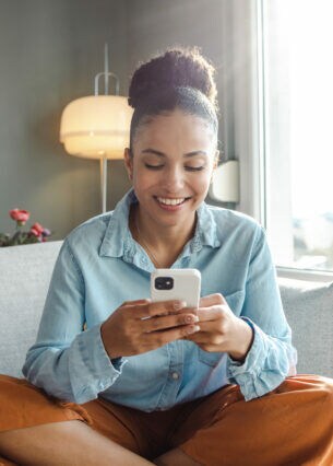 Eine Person sitzt im Schneidersitz auf einem Sofa und blickt auf ihr Smartphone, das sie in beiden Händen hält.