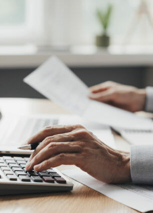 Ein Mann nutzt einen Taschenrechner und hält ein Papier in der Hand. Vor ihm steht ein Laptop auf dem Tisch.