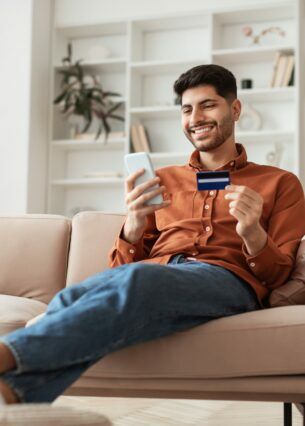 Eine Person sitzt mit ausgestreckten Beinen auf einem Sofa und hält eine Kreditkarte sowie ein Smartphone in den Händen.