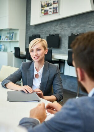 Drei Personen sitzen an einem weißen Tisch, auf dem sich ein Stiftbehälter und ein Trinkglas befinden. Und Heftmappen, auf die eine Person ihre Hand ablegt.
