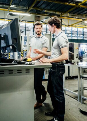 Zwei Personen in Arbeitskleidung stehen nebeneinander in einer Produktionshalle an einem Tisch, auf dem sich ein Bildschirm befindet.