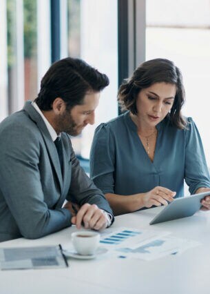 Zwei Personen in Businesskleidung sitzen an einem weißen Tisch, vor sich Kaffeetassen, ein Tablet und diverse Dokumente.