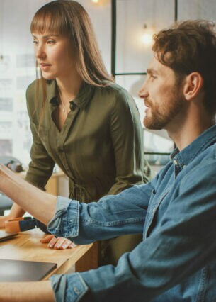 Ein Mann am Schreibtisch zeigt einer neben ihm stehenden Frau am PC-Monitor ein Porträt einer Person in einer Fotobearbeitungssoftware.