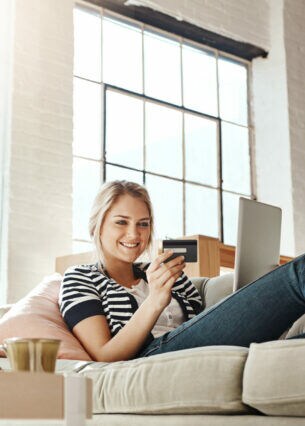 Eine Person liegt auf einem Sofa, hat ein Laptop auf ihrem Schoß und blickt lächelnd auf eine Bankkarte.