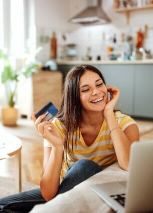 Eine Person sitzt auf dem Fußboden zwischen Couchtisch und Sofa, hält eine Kreditkarte in der Hand und blickt auf ein geöffnetes Notebook, das auf der Sofasitzfläche steht. 