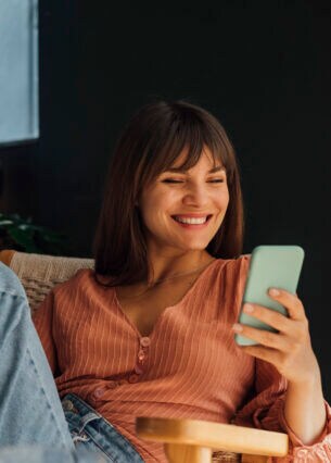 Eine Person sitzt in einem Sessel und blickt lachend auf ihr Smartphone, das sie in der Hand hält.