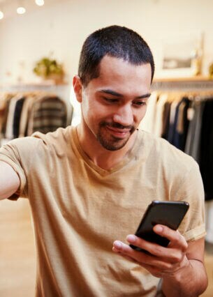 Eine Person steht in einem Bekleidungsgeschäft, greift mit einer Hand nach einem Paar Sneaker und hält in der anderen Hand ein Smartphone.