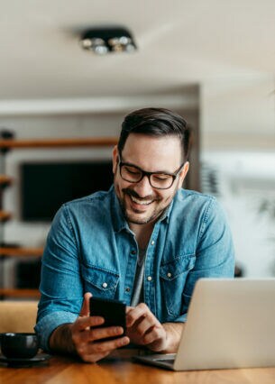 Eine Person sitzt an einem Tisch und blickt auf ein Smartphone, das sie in den Händen hält. Vor ihr stehen ein geöffnetes Laptop und eine Kaffeetasse.