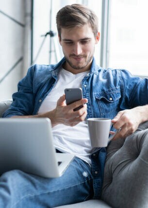 Eine Person sitzt mit ausgestreckten Beinen auf einem Sofa, hat auf dem Schoß ein geöffnetes Laptop liegen und hält in ein Smartphone sowie eine Tasse in den Händen. 