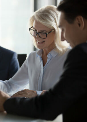 Drei Personen in Businesskleidung sitzen an einem Tisch, auf dem sich ein aufgeklappter Laptop und Papierunterlagen befinden.