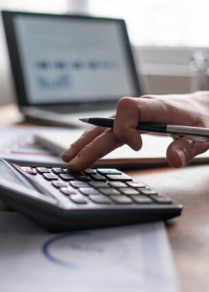 Eine Hand hält einen Stift und tippt etwas auf einem Taschenrechner. Im Hintergrund ist ein Laptop mit Diagrammen zu sehen.