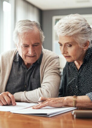 Eine ältere Frau und ein älterer Mann sitzen am Tisch. Sie schauen sich aufmerksam Dokumente an, die vor ihnen liegen.