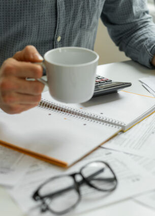 Eine Person am Tisch hält eine Tasse und einen Kugelschreiber in Händen. Auf dem Tisch liegen Dokumente, ein Taschenrechner und eine Brille.