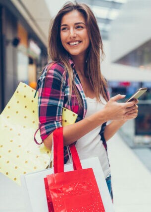 Eine lächelnde Person steht in einer Shoppingmall und hat mehrere Tüten über die Schulter gehängt und hält ein Smartphone in den Händen.