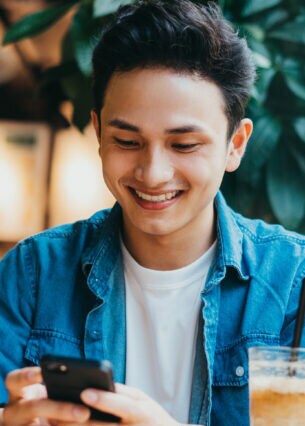 Eine Person sitzt mit einem Café vor sich am Tisch und blickt auf das Smartphone, das sie in den Händen hält.