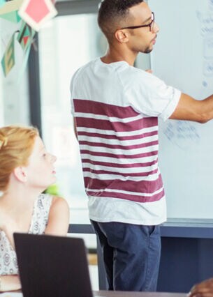 Mann steht mit einem Stift in der Hand an einem Whiteboard und zeigt seinen drei Arbeitskolleg:innen seine Notizen.