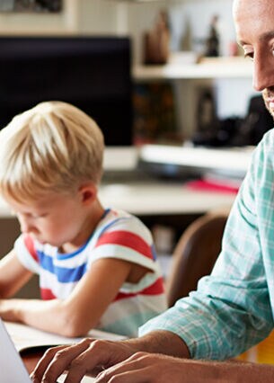 Blogbild: Person arbeitet am laptop während ein Kind daneben Hausaufgaben macht