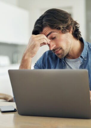 Ein Mann sitzt vor einem Laptop an einem Tisch, hat die Augen geschlossen und eine Hand an seine Stirn gelegt.