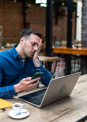Ein Mann sitzt an einem Tisch vor einem Laptop und hält ein Smartphone in der Hand. Er hat die Augen geschlossen und legt eine Hand über sein Gesicht.