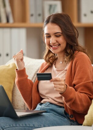 Eine Person sitzt lächelnd auf einem Sofa, einen Laptop auf den Beinen und eine Kreditkarte in der Hand und ballt eine Faust.