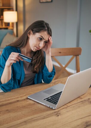 Eine Frau sitzt mit gerunzelter Stirn an einem Tisch vor einem Laptop. In der Hand hält sie eine blaue Karte.