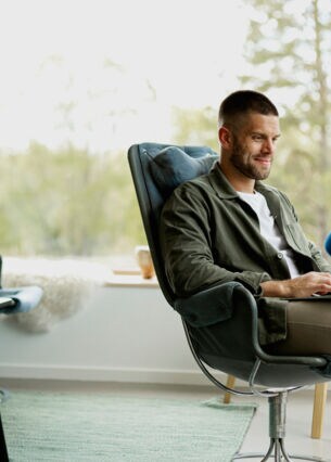 Eine Person sitzt vor einer großen Fensterfront in einem Sessel mit einem Laptop auf dem Schoß.