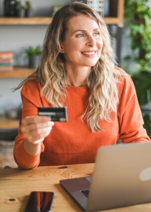 Eine Frau sitzt lächelnd an einem Tisch. Vor ihr steht ein Laptop und in der Hand hält sie eine Kreditkarte.