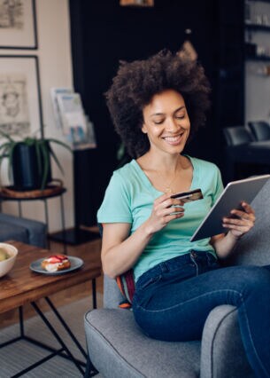Person sitzt auf einem Sessel und schaut auf ihr Tablet und auf ihre Kreditkarte