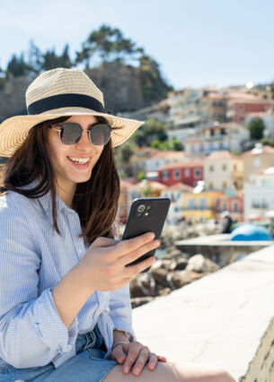 Person schaut auf ihr Handy während sie auf einer Mauer sitzt. Hinter ihr ist das Meer und bunte Häuser
